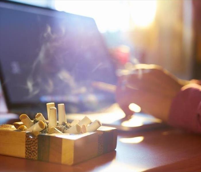 Close up of ashtray full of cigarette, with man in background working on laptop computer and smoking indoors on early morning